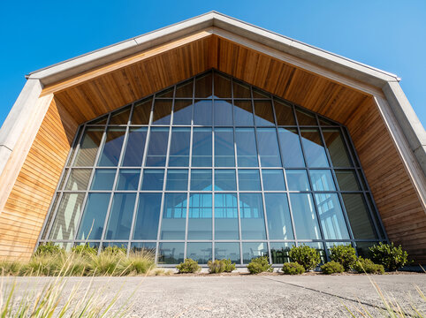 Gable end view of a building with a large glass curtain wall, wooden soffit, and concrete foreground under a clear blue sky