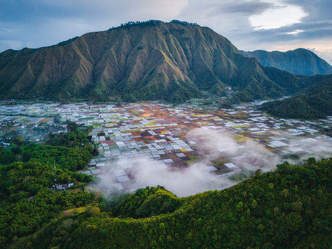 Aerial view of the Sembalun agricultural basin with colorful patchwork fields at the foothills of Mount Rinjani, featuring low-hanging clouds in Sembalun, West Nusa Tenggara, Indonesia.