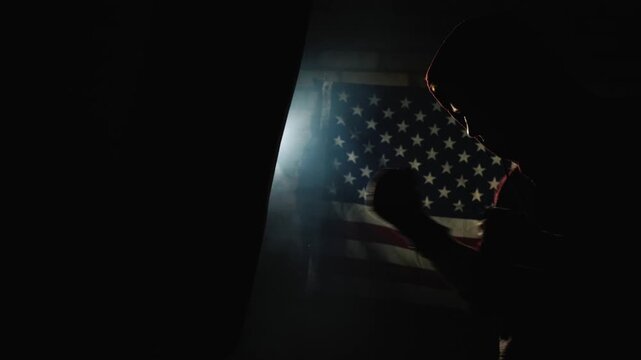 Boxer training on punching bag in dark gym with american flag in background.