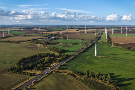 Aerial view of a vast wind farm with numerous wind turbines situated in green agricultural fields alongside a highway with moving trucks in Uckermark, Brandenburg, Germany.
