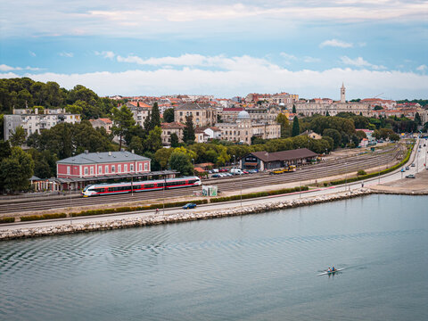 Aerial view of a train station situated along the coastline with a red and white train on the tracks, nearby buildings, and a small boat rowing on the calm sea in Buzet, Istria County, Croatia.