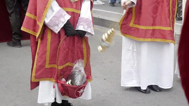 Acolytes in traditional red robes holding incense burner and basket during religious street procession.