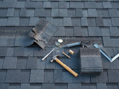 Roofing tools and dark grey shingles on a damaged roof ready for repair and replacement.