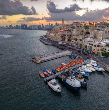 Aerial view of the Old port of Jaffa with fishing boats and yachts moored at the pier, historic stone buildings, and the modern Tel Aviv skyline at sunset, Tel Aviv-Yafo, Tel Aviv District, Israel.
