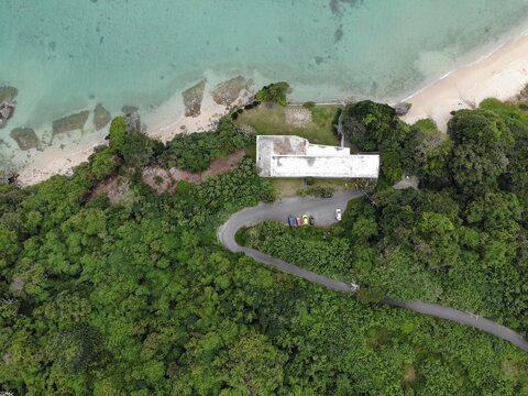 Aerial view of the Hyakume coastline with a building in lush greenery, a winding road with parked cars, and a sandy beach meeting the emerald sea in Nanjo, Okinawa, Japan.