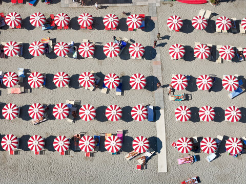 Aerial view of rows of red and white striped beach umbrellas on the sandy beach with people relaxing on lounge chairs in Bergeggi, Liguria, Italy.