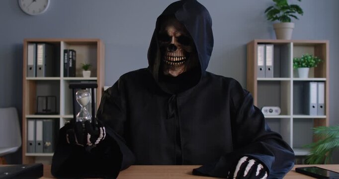 Death in black hooded Grim Reaper cloak sitting at office desk takes sand glass timepiece in skeleton hand and starts beating time reminding that clock is ticking and work deadline is approaching