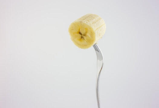 A single peeled banana slice is impaled on the tines of a metal fork, set against a plain white studio background in a still life shot.
