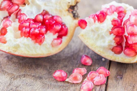 A split ripe pomegranate with bright red seeds, some scattered on a rustic wooden surface, close-up shot of fresh fruit.