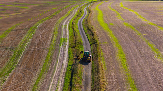 Aerial view of a tractor trimming hedges along a narrow country road winding through agricultural fields at Pepperbox Hill. Salisbury, England, United Kingdom.