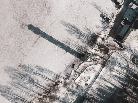 Aerial view of Majak Jary Cimrmana casting a long shadow over a snow-covered landscape with nearby buildings and trees in Korenov, Liberecky kraj, Czechia.