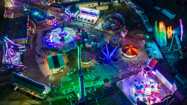 Aerial view of a vibrant fairground at night featuring illuminated amusement rides, spinning attractions, and visitors walking through the park in Vilnius, Vilnius County, Lithuania.