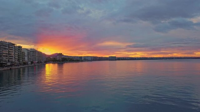 THESSALONIKI, GREECE: Cinematic aerial video flying forward over the calm sea of Thermaikos gulf, revealing the city's coastal skyline and waterfront buildings under a dramatic, colorful pink and oran