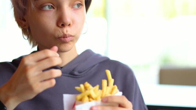 This engaging video shows a colorful-haired teenage girl eating French fries in a fast-food cafe, capturing her relaxed mood and the simple pleasure of a casual meal.