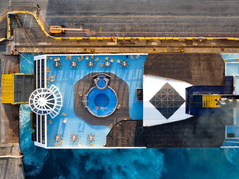 Aerial view of the deck of a ferry to Corsica, featuring a circular blue swimming pool, outdoor seating with tables and chairs, and the ship docked at the pier in Vado Ligure, Liguria, Italy.
