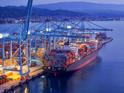 Aerial view of APM Terminals Vado Ligure container port with a large cargo ship docked, colorful shipping containers, and tall gantry cranes at dusk in Vado Ligure, Liguria, Italy.