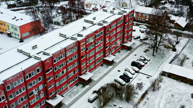 Aerial view of a red apartment building surrounded by snow-covered grounds, parking lot with cars, and a playground in Latvija.