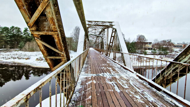 Aerial view of an old, rusty metal truss bridge covered in snow spanning a river in a winter landscape in Latvia.