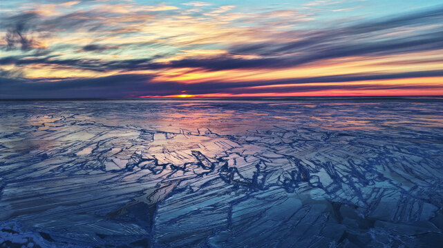 Aerial view of a vast expanse of cracked ice on a frozen lake at sunset with a vibrant, colorful sky in Latvia.