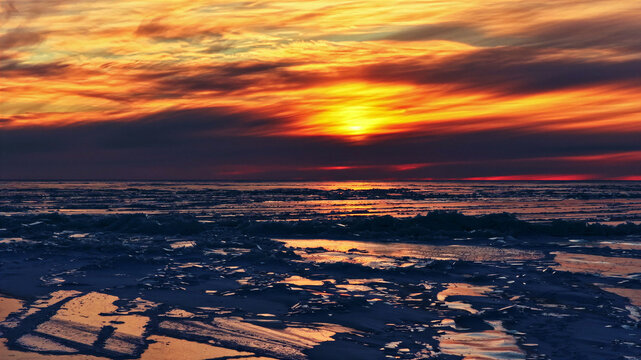 Aerial view of a frozen landscape with textured ice and snow under a dramatic sunset sky with vibrant orange and yellow clouds in Latvia.