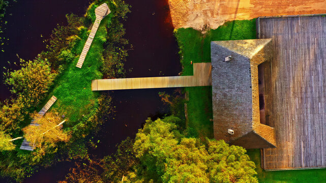 Aerial view of a wooden building with a large deck and a wooden walkway extending over a dark pond surrounded by lush green trees and vegetation in Latvia.