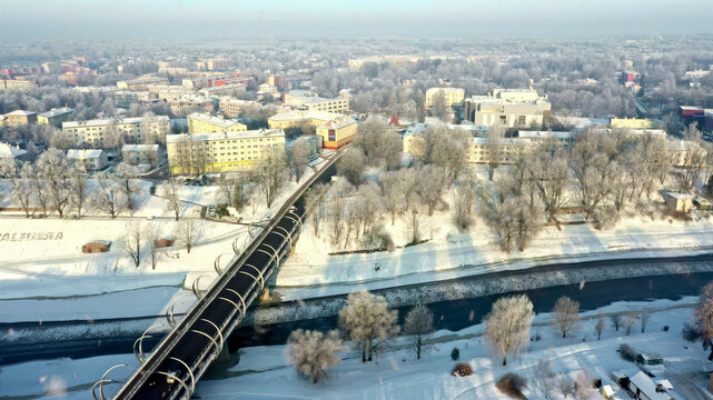 Aerial view of a modern bridge spanning a river in a snow-covered town with frost-covered trees and residential buildings in Latvija.