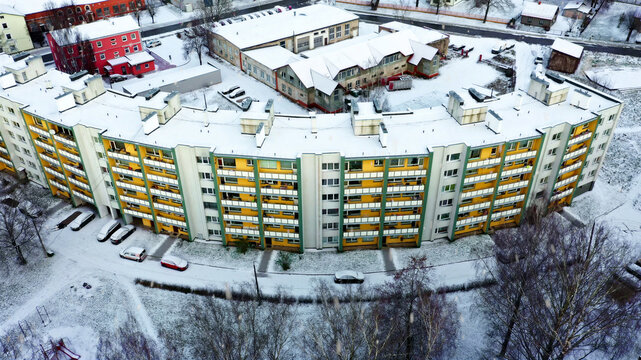 Aerial view of a curved multi-story apartment building with yellow and green facades covered in a light dusting of snow, surrounded by trees and a parking lot in Latvia.