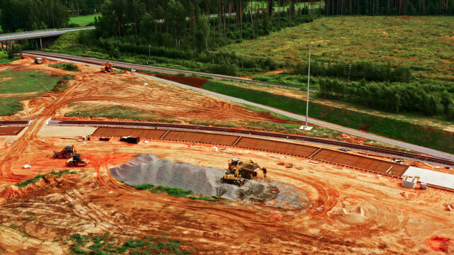 Aerial view of a railway construction site with heavy machinery, excavators, and piles of gravel near a forest and railway tracks in Latvia.