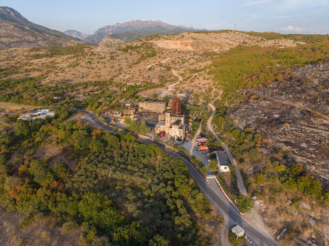 Aerial view of an industrial facility nestled in a mountainous landscape with olive groves, a winding road, and signs of recent vegetation fire on the hillside in Boks, Qarku i Shkodres, Albania.