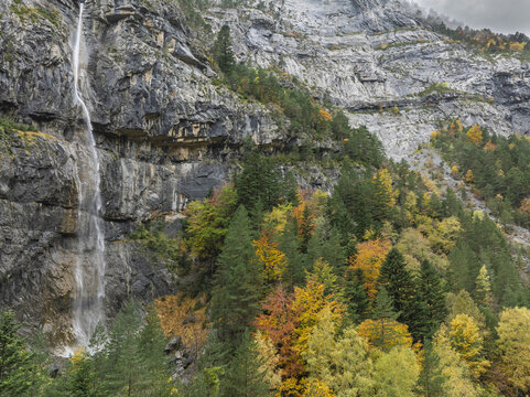 Aerial view of a waterfall cascading down a steep rocky cliff face in the Pyrenees, surrounded by a vibrant autumn forest with colorful trees in Spain.