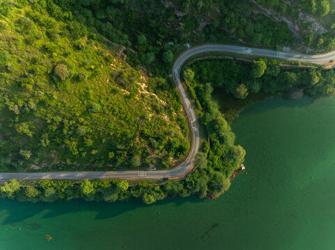 Aerial view of a winding road along the edge of a green lake surrounded by lush hills in Udaipur, Rajasthan, India.