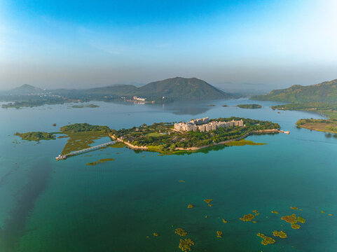 Aerial view of the Taj Lake Palace situated on an island in Lake Pichola, surrounded by calm turquoise waters and distant hills in Udaipur, Rajasthan, India.