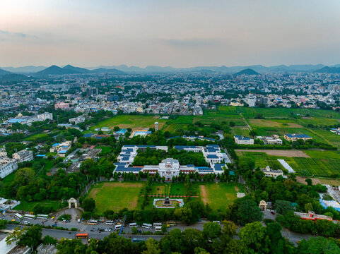 Aerial view of a large institutional building complex surrounded by lush green lawns, trees, and agricultural fields with the cityscape of Udaipur, Rajasthan, India.