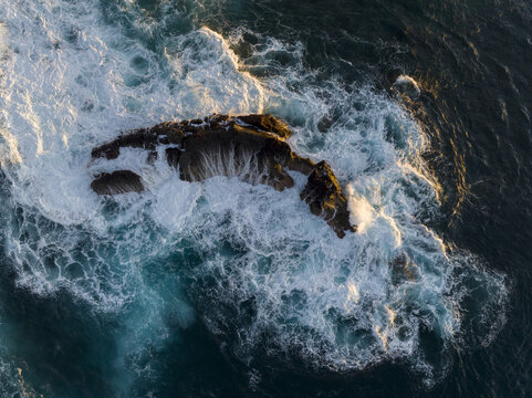 Aerial view of a rugged volcanic rock formation surrounded by crashing white waves and deep blue Atlantic ocean water during golden hour in Lanzarote, Canarias, Spain.