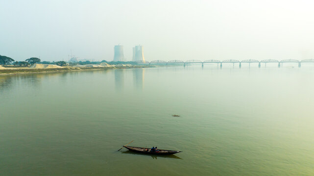 Aerial view of a small wooden boat floating on the calm Padma River with the distant Hardinge Bridge and cooling towers of the Rooppur Nuclear Power Plant in Pabna, Rajshahi Division, Bangladesh.