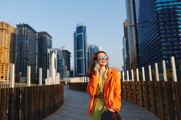 Small figure in a bright orange blazer stands on a wooden boardwalk amid towering city skyscrapers, smiling while talking on a phone. Concept for urban careers or recruitment campaigns. © SHOTPRIME STUDIO