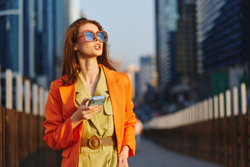 Sunlit profile portrait of a stylish woman in an orange blazer, holding a phone, against a modern cityscape. Emphasizes confident stance, warm light, and fashion-forward urban wellness vibe. © SHOTPRIME STUDIO