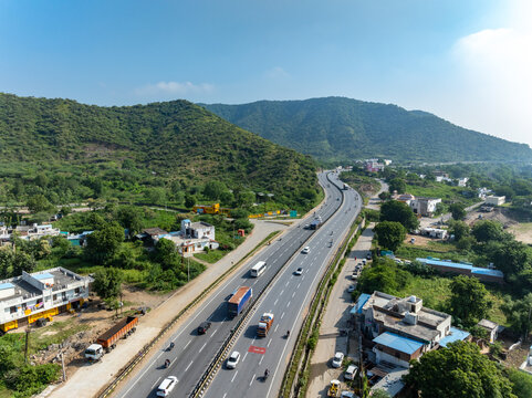 Aerial view of a multi-lane highway with cars and trucks winding through green hills and residential buildings in Udaipur, Rajasthan, India.