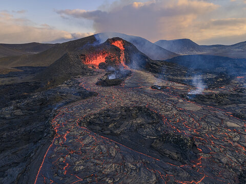 Aerial view of a volcanic eruption with glowing lava flowing from a crater across a rugged black basalt landscape in Iceland.