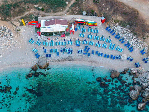 Aerial view of Riserva Naturale Orientata dello Zingaro with turquoise water, pebble beach, colorful kayaks, and rows of sun loungers in San Vito Lo Capo, Sicilia, Italy.