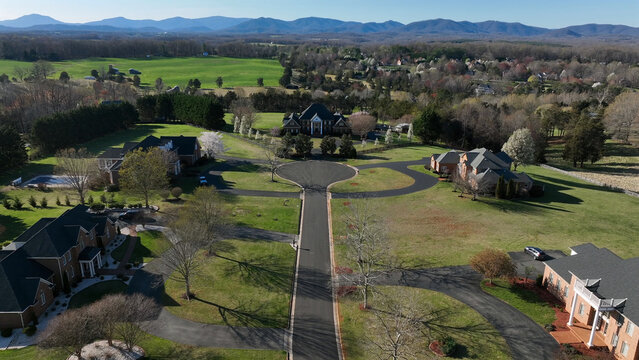 Aerial view of a suburban residential neighborhood with large houses, manicured lawns, winding driveways, and distant mountains under a clear blue sky in Forest, Virginia, United States.