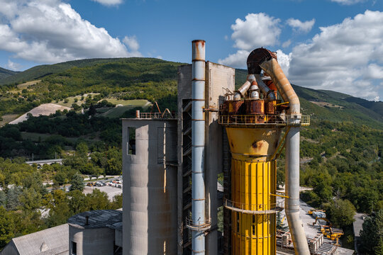 Aerial view of the Sacci S.P.A. cement plant featuring a large yellow industrial silo and concrete structures set against a backdrop of green hills, Castelraimondo, Marche, Italy.