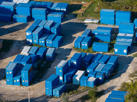 Aerial view of numerous blue shipping containers arranged in an industrial storage yard with gravel ground and shadows cast by the afternoon sun in Nera Montoro, Umbria, Italy.