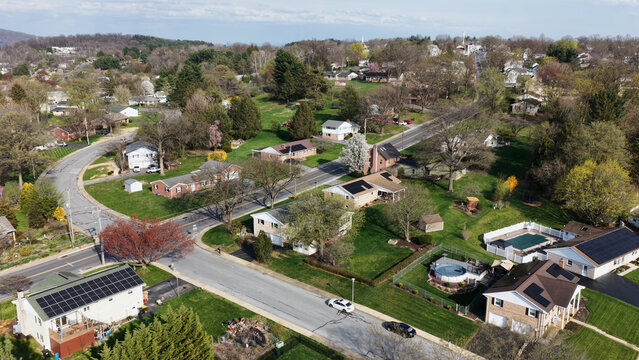 Aerial view of a suburban neighborhood featuring residential houses with solar panels, green lawns, swimming pools, and tree-lined streets in Akron, Pennsylvania, United States.
