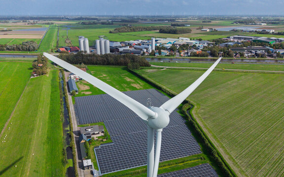Aerial view of a large wind turbine standing next to a vast solar panel farm in the green rural landscape of 't Zand, North Holland, Netherlands.