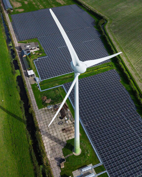 Aerial view of a wind turbine standing next to a large solar panel farm in a rural landscape with green fields and a small house in 't Zand, North Holland, Netherlands.