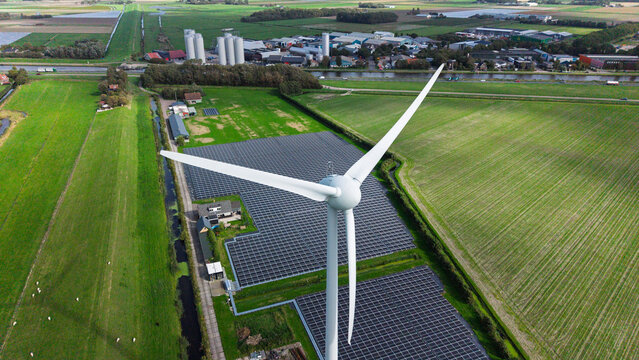 Aerial view of a wind turbine and a large solar panel array situated in green fields near a canal and industrial buildings in 't Zand, North Holland, Netherlands.