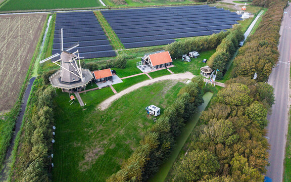Aerial view of Molen De Hoop windmill surrounded by a large solar panel field, green meadows, and a small building with a camper van in 't Zand, North Holland, Netherlands.