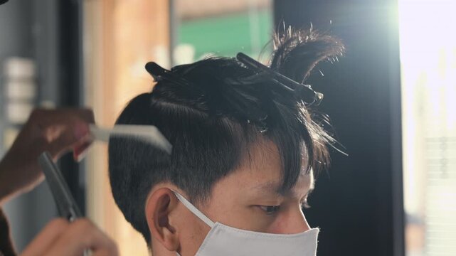 A barber cuts a seated customer&rsquo;s dark hair with scissors during a haircut. The barber holds a comb near the head while the customer wears a face mask and protective cape inside the salon.
