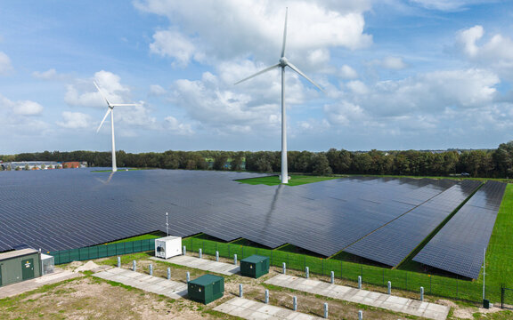 Aerial view of a large solar farm with rows of photovoltaic panels and wind turbines set in a green landscape in Warmenhuizen, North Holland, Netherlands.